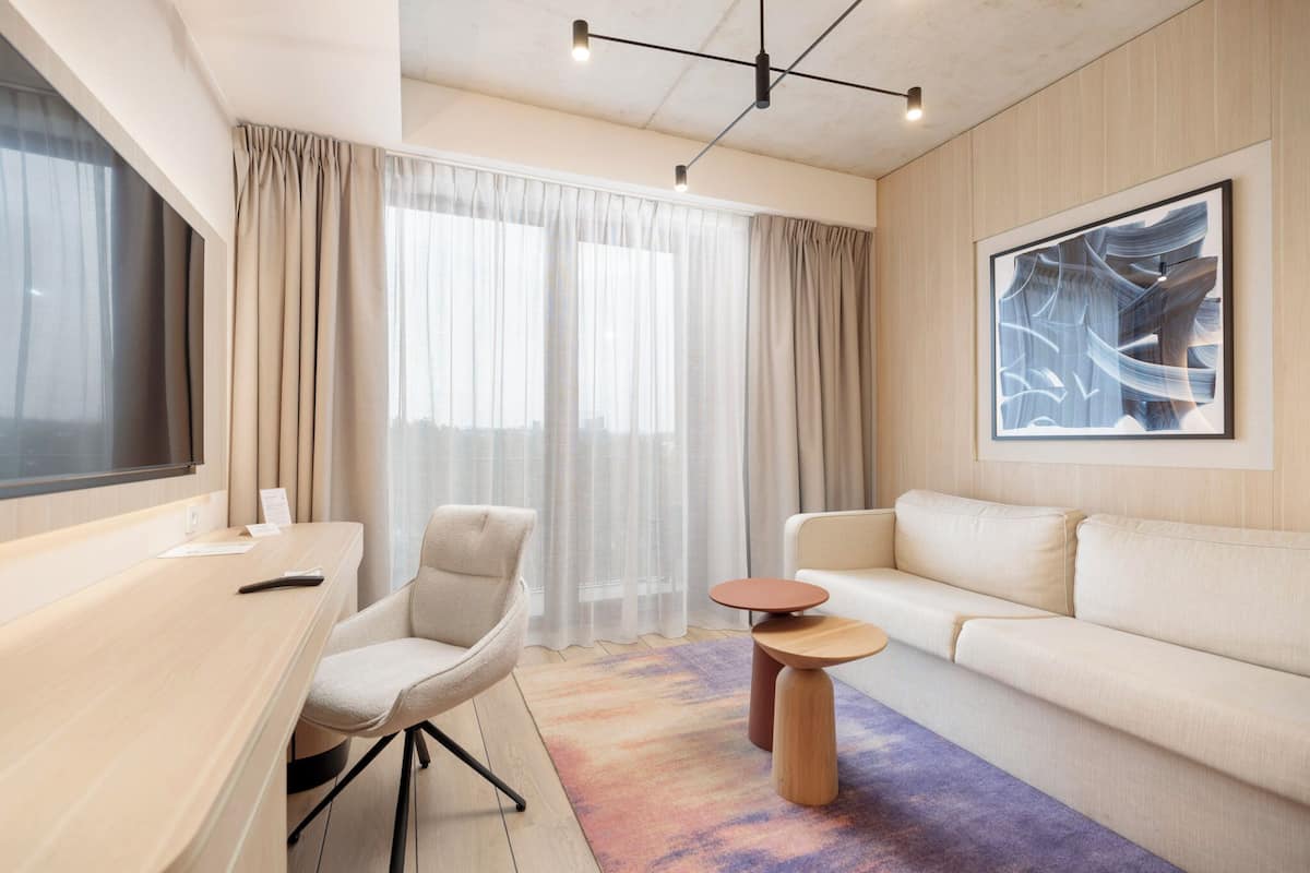 A modern hotel room with a large window, a sofa, a desk, and a coffee table.