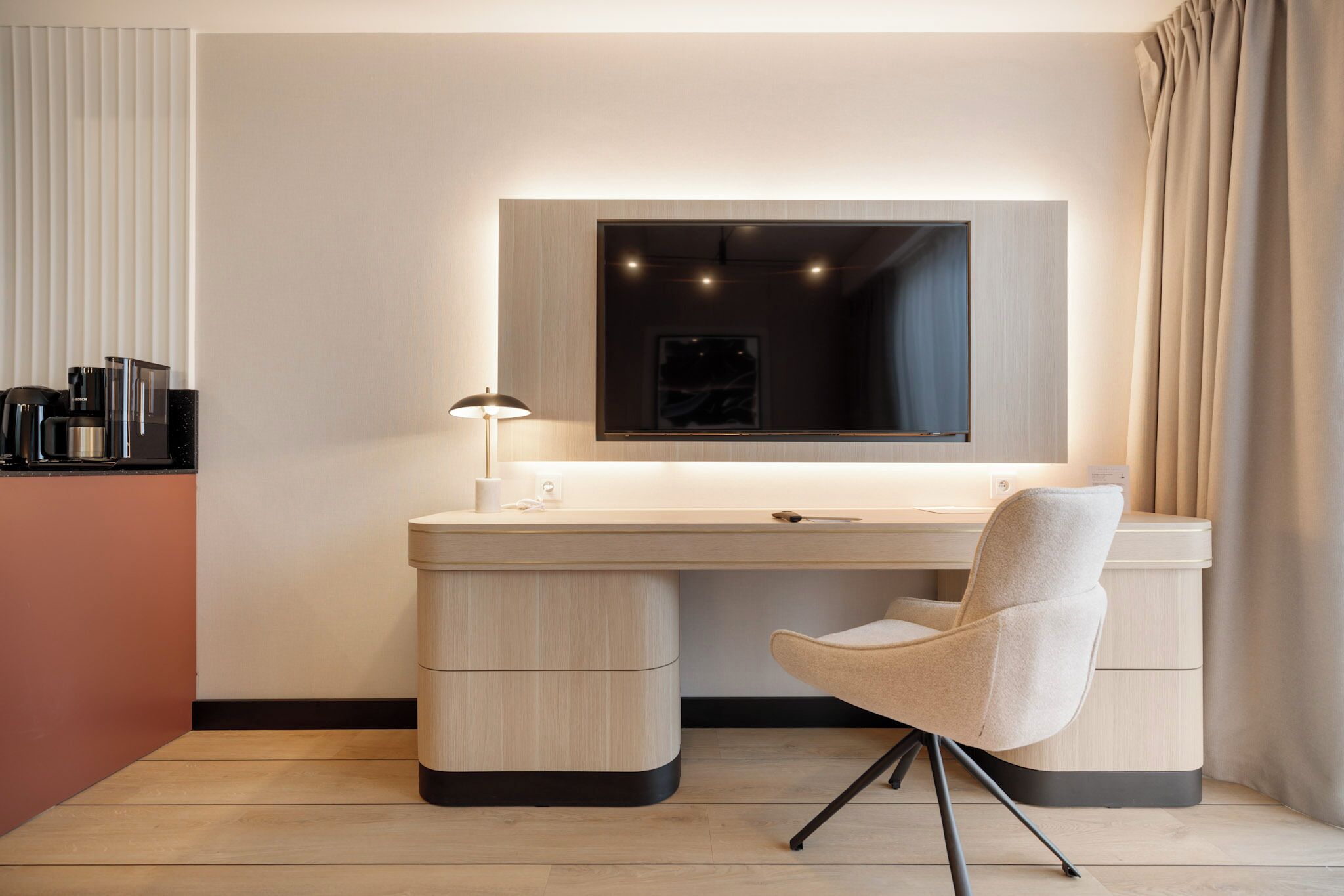A modern hotel room with a wooden desk, a chair, a large mirror, and a bedside table.