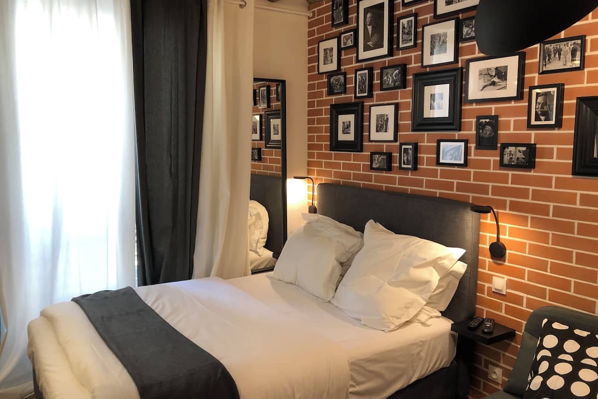 A hotel room with a brick-patterned wall, a large bed with white bedding, a nightstand with a lamp, and a chair with a polka dot cushion.
