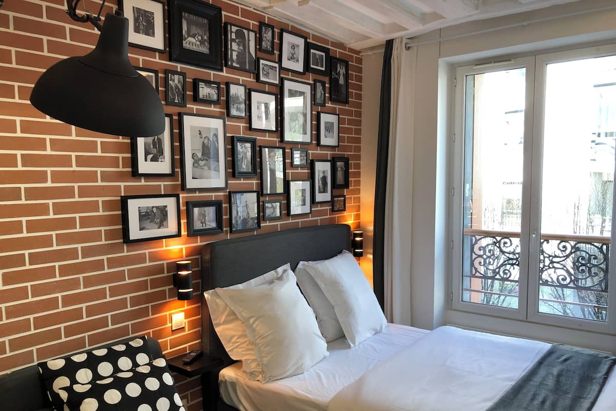 A hotel room with a brick wall, a large bed with white bedding, a black polka dot pillow, a black pendant lamp, and framed pictures on the wall.