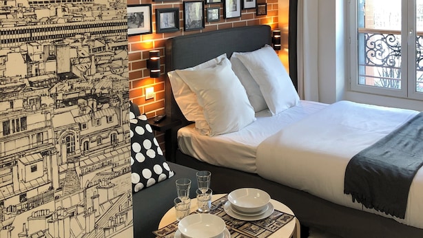 A hotel room with a bed, a nightstand with a lamp, a wall with framed pictures, and a window with a view of the city. - Résidence Labyrinthe (Paris)