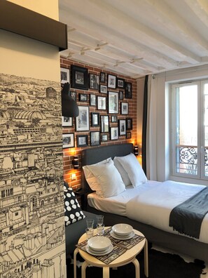 A hotel room with a bed, a nightstand with a lamp, a wall with framed pictures, and a window with a view of the city. - Résidence Labyrinthe (Paris)