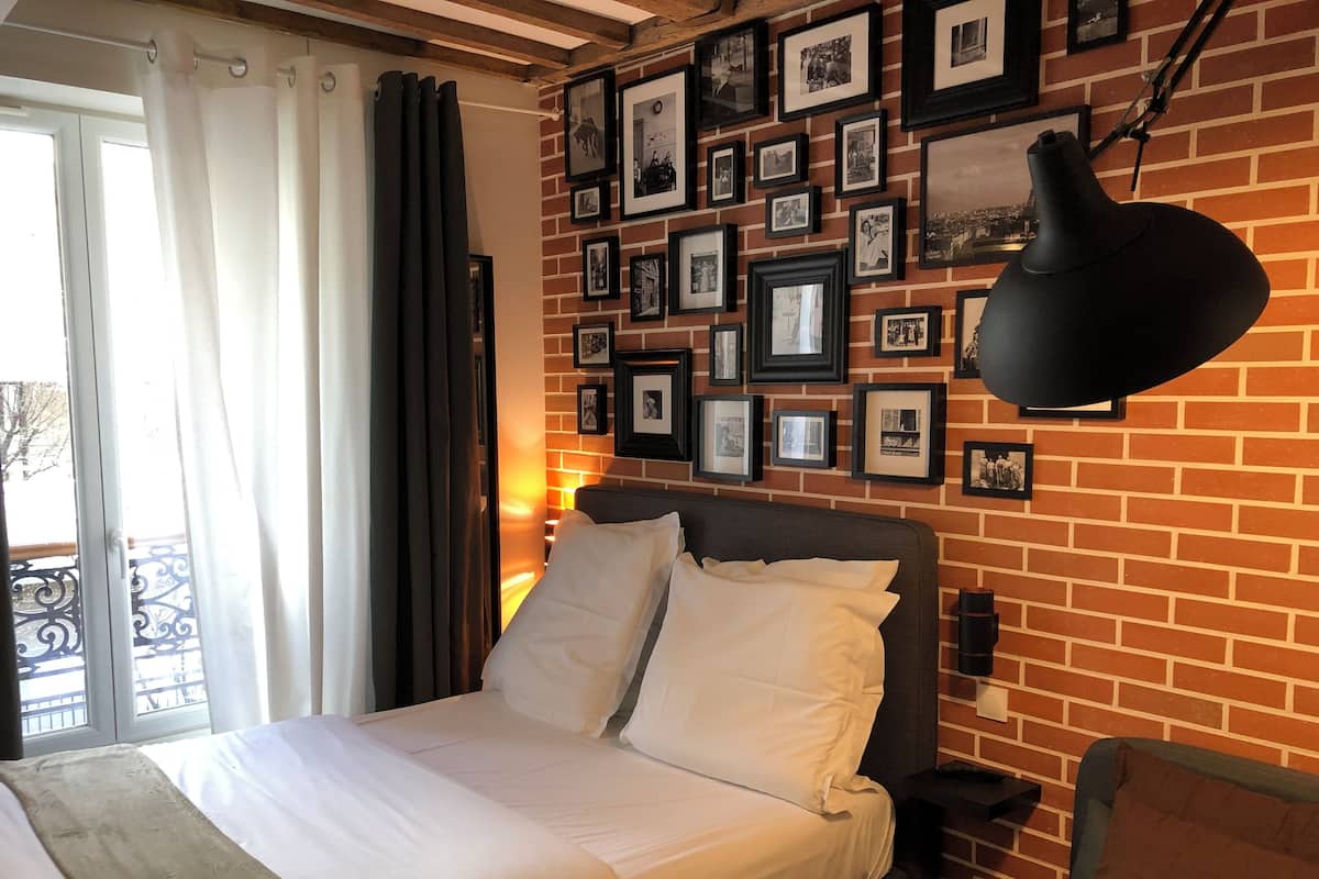A hotel room with a brick wall, a bed with white linens, a black lamp, and a balcony with a view.