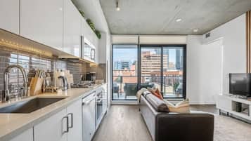 A modern apartment with a kitchen, a living area with a sofa and a TV, and large windows offering a city view.