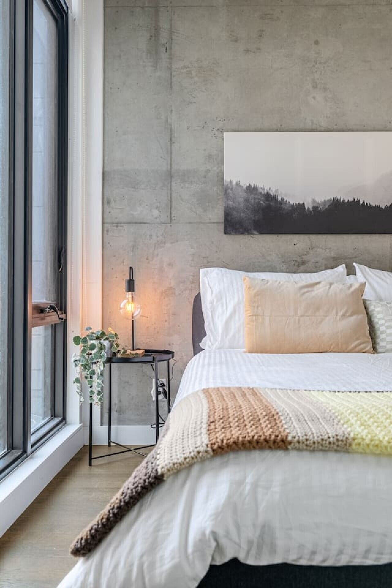 A bedroom with a large bed, a wall-mounted painting, a bedside table with a lamp, and a window with a view.