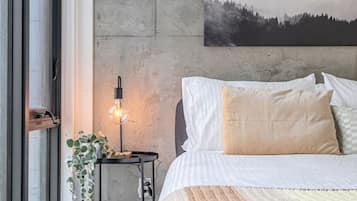A bedroom with a large bed, a wall-mounted painting, a bedside table with a lamp, and a window with a view.