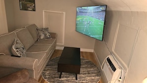 Ein Wohnzimmer mit einem an der Wand befestigten Flachbildfernseher, einem grauen Sofa mit gemusterten Kissen, einem schwarzen Couchtisch und einem Teppich mit Muster.