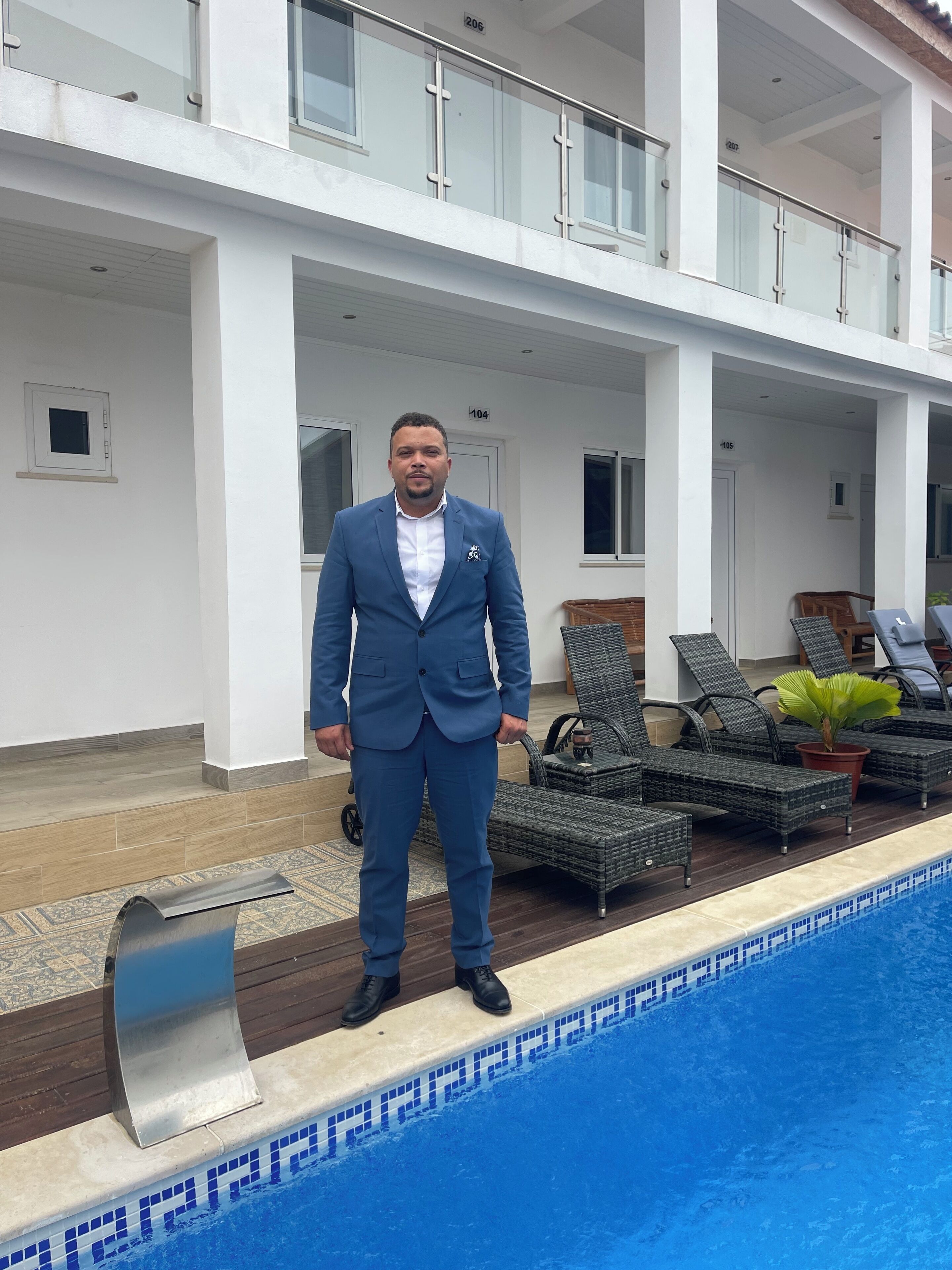 Un homme en costume bleu se tient près d’une piscine, avec des chaises longues à proximité et un bâtiment blanc en arrière-plan.