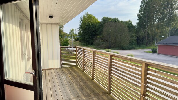A wooden deck with a railing, a glass door, and a view of a road and trees. - Beautiful house with backyard  (Partille)