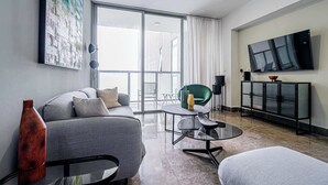 Un salon moderne avec un canapé, une table basse en verre, un fauteuil vert, un téléviseur et une grande fenêtre.