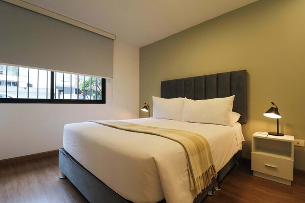 A modern hotel room with a large bed, a nightstand with a lamp, and a window with blinds.