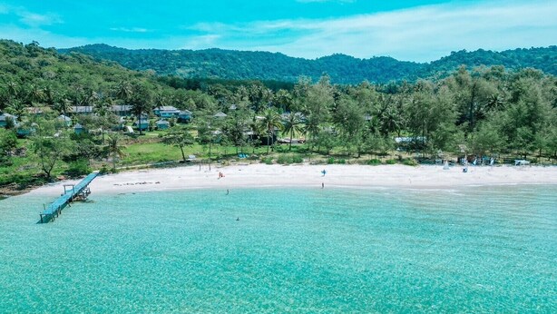 Ein Strand mit klarem türkisblauem Wasser, einer Holzsteg und im Hintergrund üppige, grüne Hügel sowie Häuser.