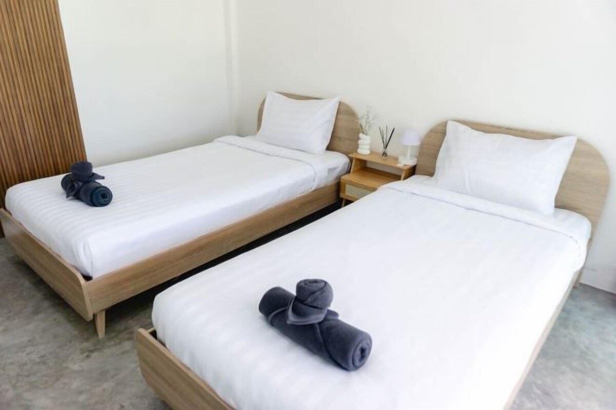 Habitación de hotel con dos camas individuales, cada una con estructura de madera y ropa de cama blanca. Hay una mesita de noche pequeña con una lámpara y un objeto decorativo.
