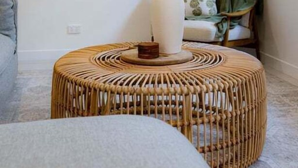 A room with a rattan ottoman, a framed landscape painting, and a window with curtains.