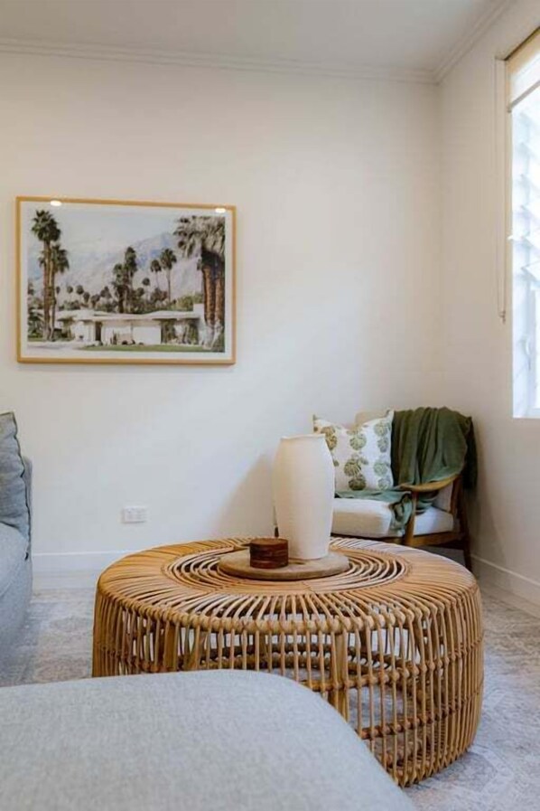 A room with a rattan ottoman, a framed landscape painting, and a window with curtains.