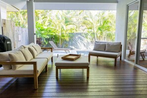 A sunlit room with a wooden floor, a beige sofa, a wooden coffee table, and a view of lush greenery through large windows. - Waves | 3 Bedroom Holiday Home (Palm Cove)