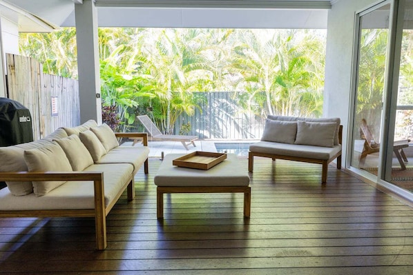 A sunlit room with a wooden floor, a beige sofa, a wooden coffee table, and a view of lush greenery through large windows.