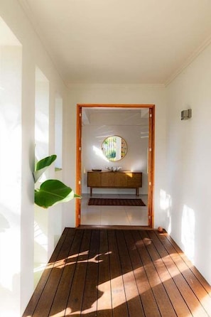A hallway with a wooden floor leading to a room with a wooden cabinet and a circular mirror on the wall. - Waves | 3 Bedroom Holiday Home (Palm Cove)