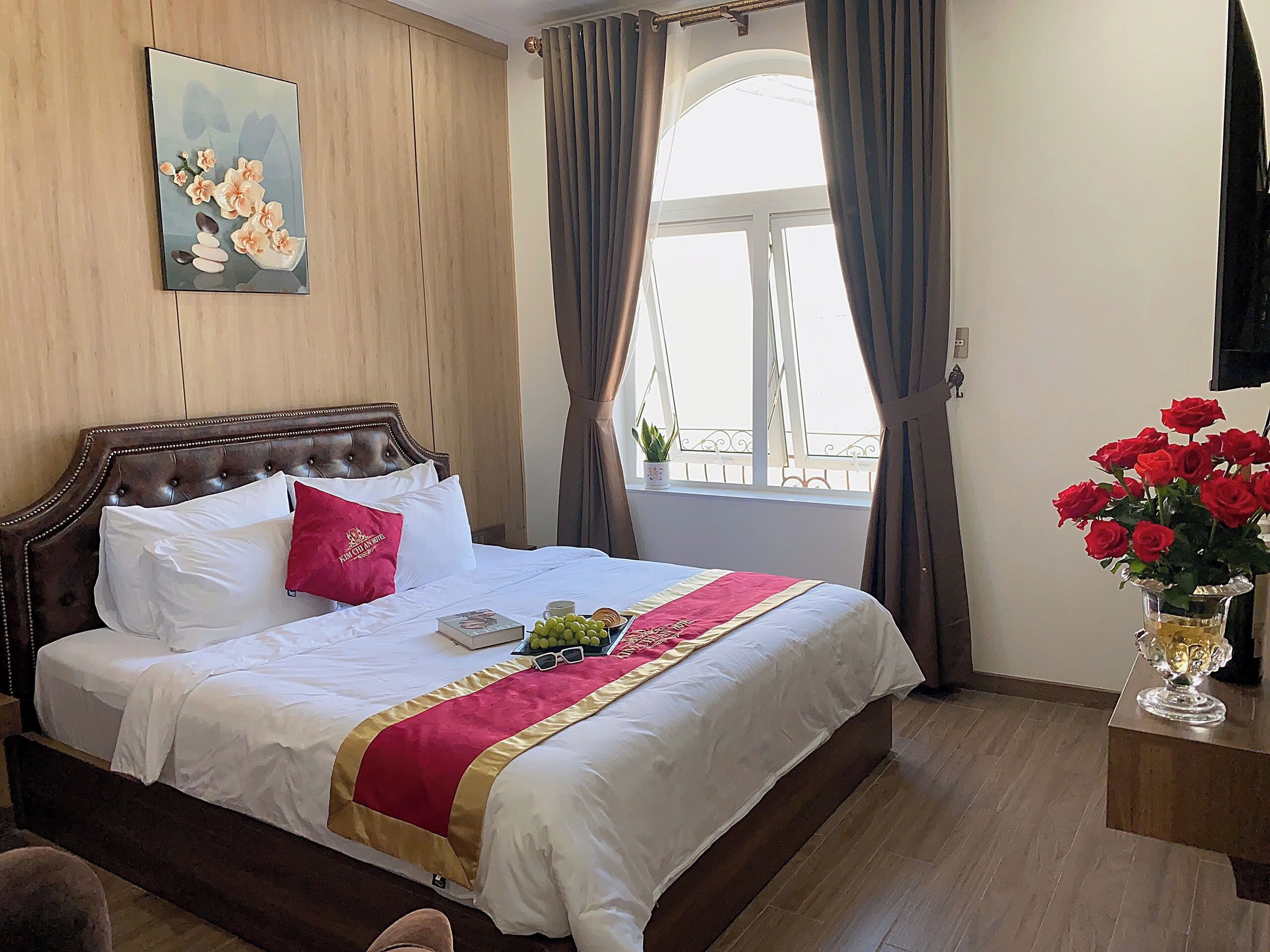 A hotel room with a large bed, a window with curtains, a vase with red flowers, and a painting on the wall.
