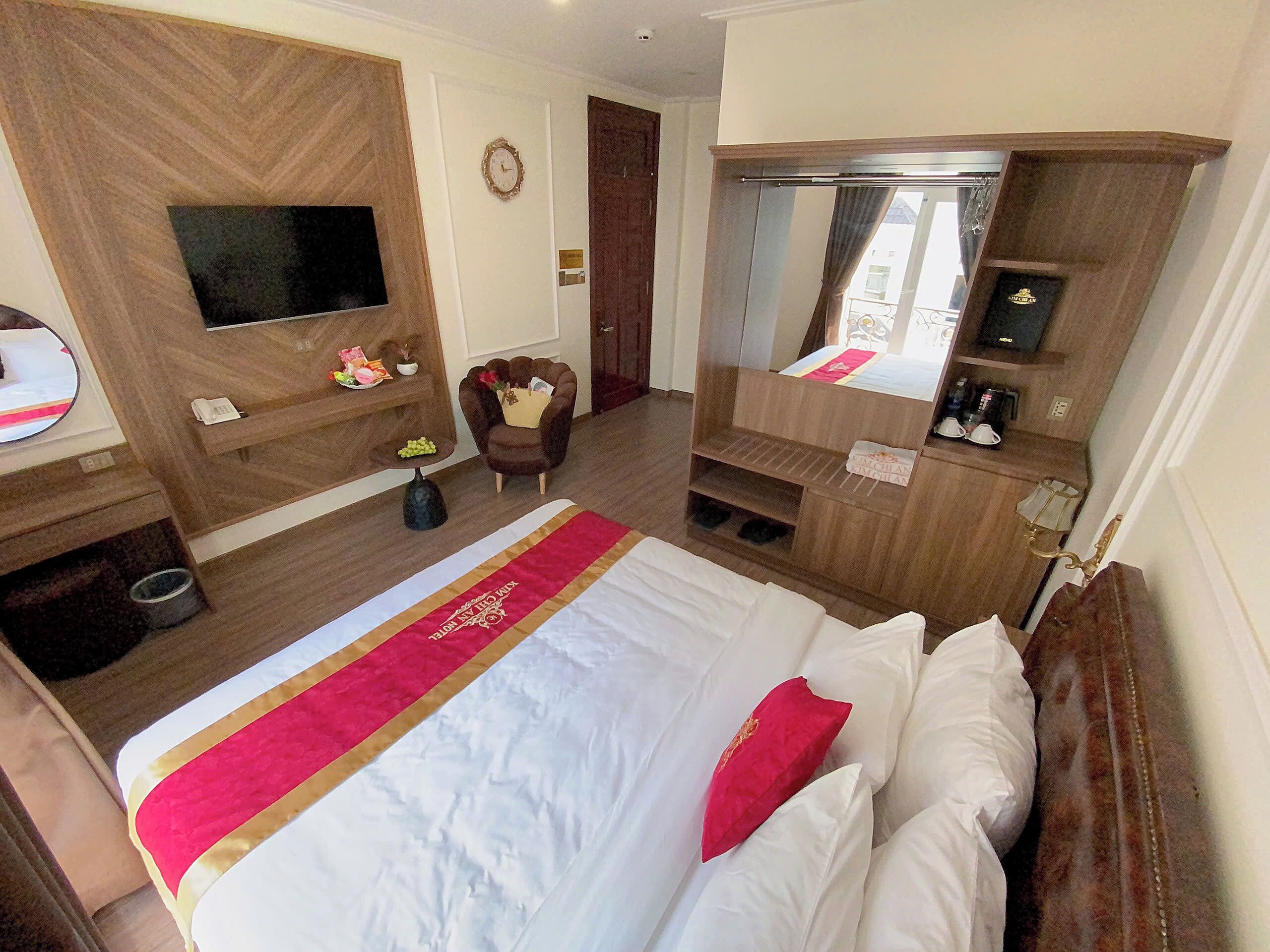 A hotel room with a large bed, a TV, a desk with a phone, a chair, a mirror, and a wardrobe.