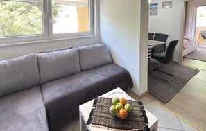Uma sala de estar moderna com um sofá cinza, uma área de jantar com mesa e cadeiras, e vista para o exterior pela janela.