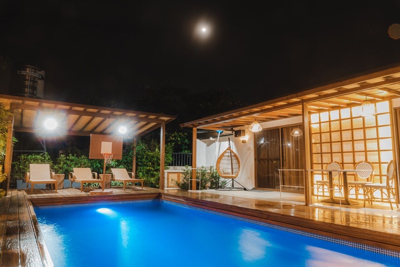 A nighttime swimming pool area with a basketball hoop, lounge chairs, and a covered seating area with tables and chairs.