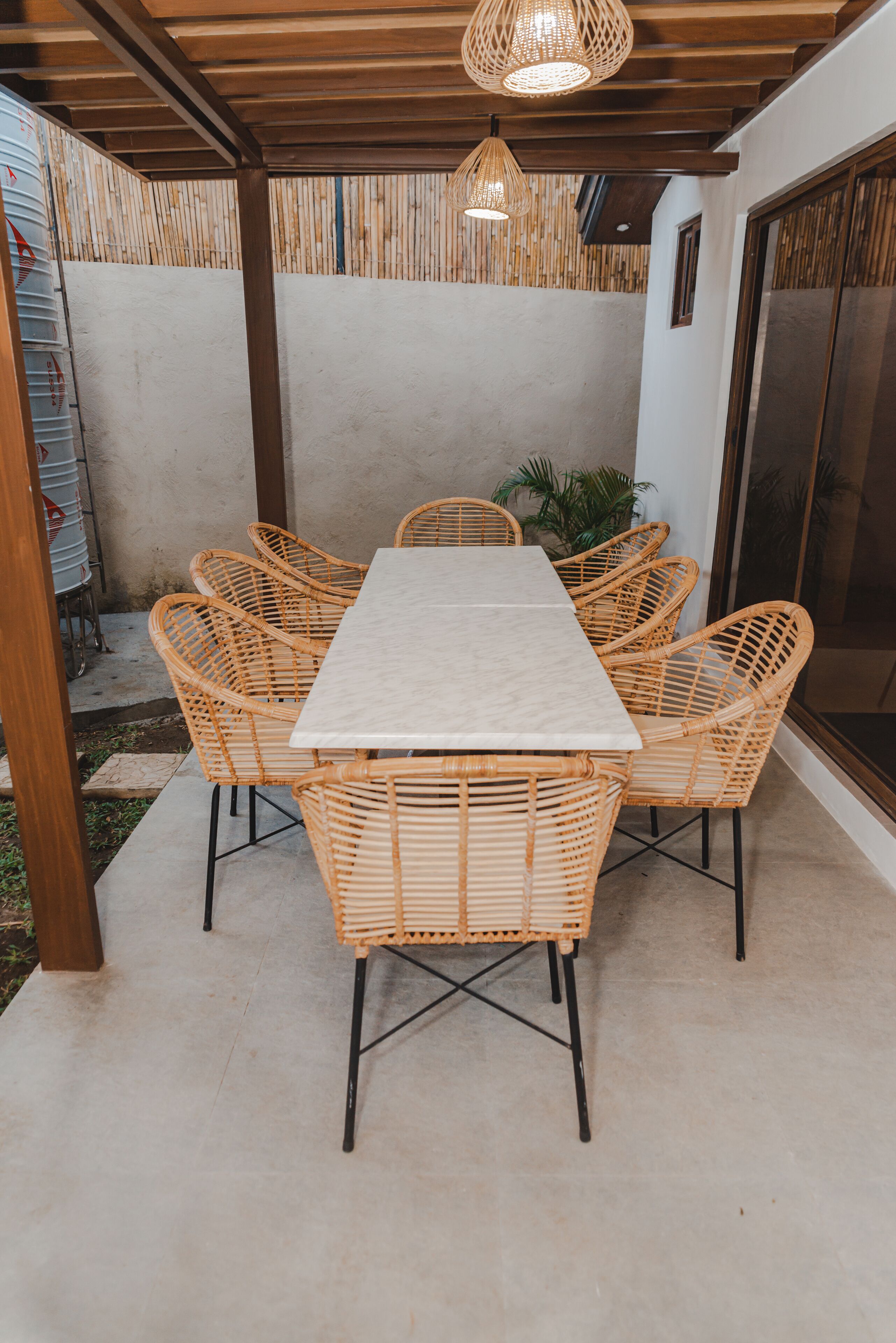 An outdoor patio with a rectangular table surrounded by wicker chairs, a hanging light fixture, and a concrete floor.