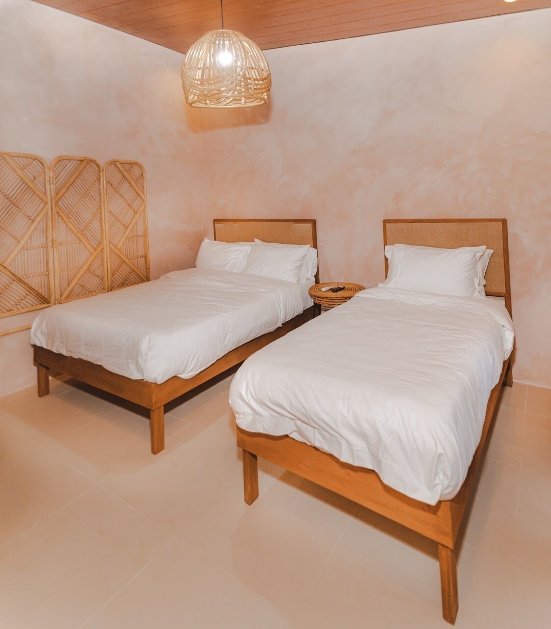 A bedroom with two wooden beds, white bedding, a woven wall decoration, and a hanging light fixture.