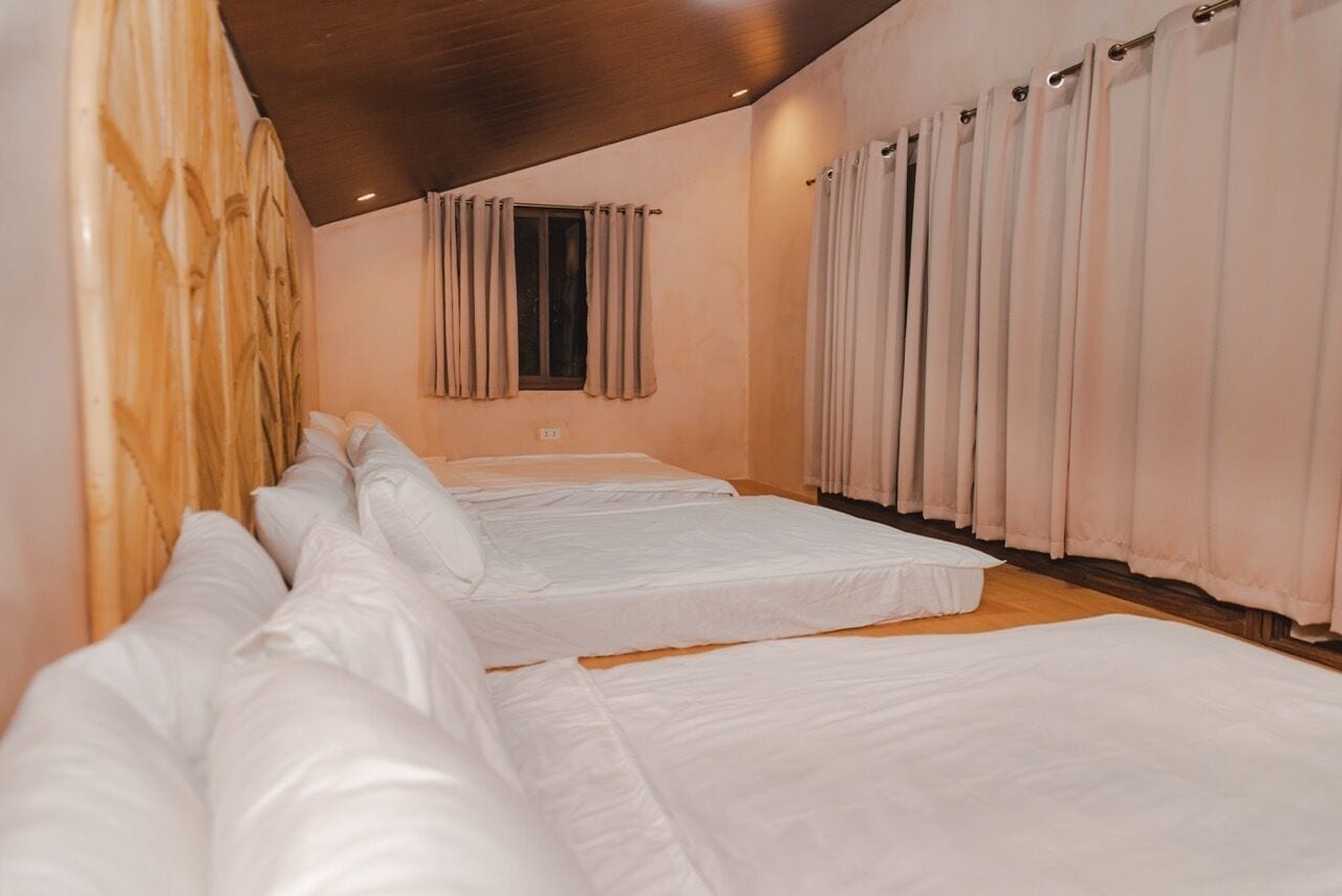 Une chambre d’hôtel avec trois lits disposés en rangée, une tête de lit en bois, une fenêtre avec des rideaux et un plafond doté d’un éclairage encastré.