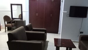 Una habitación con un armario marrón oscuro, una mesita, dos sillones, una silla, un escritorio y un televisor de pantalla plana montado en la pared.