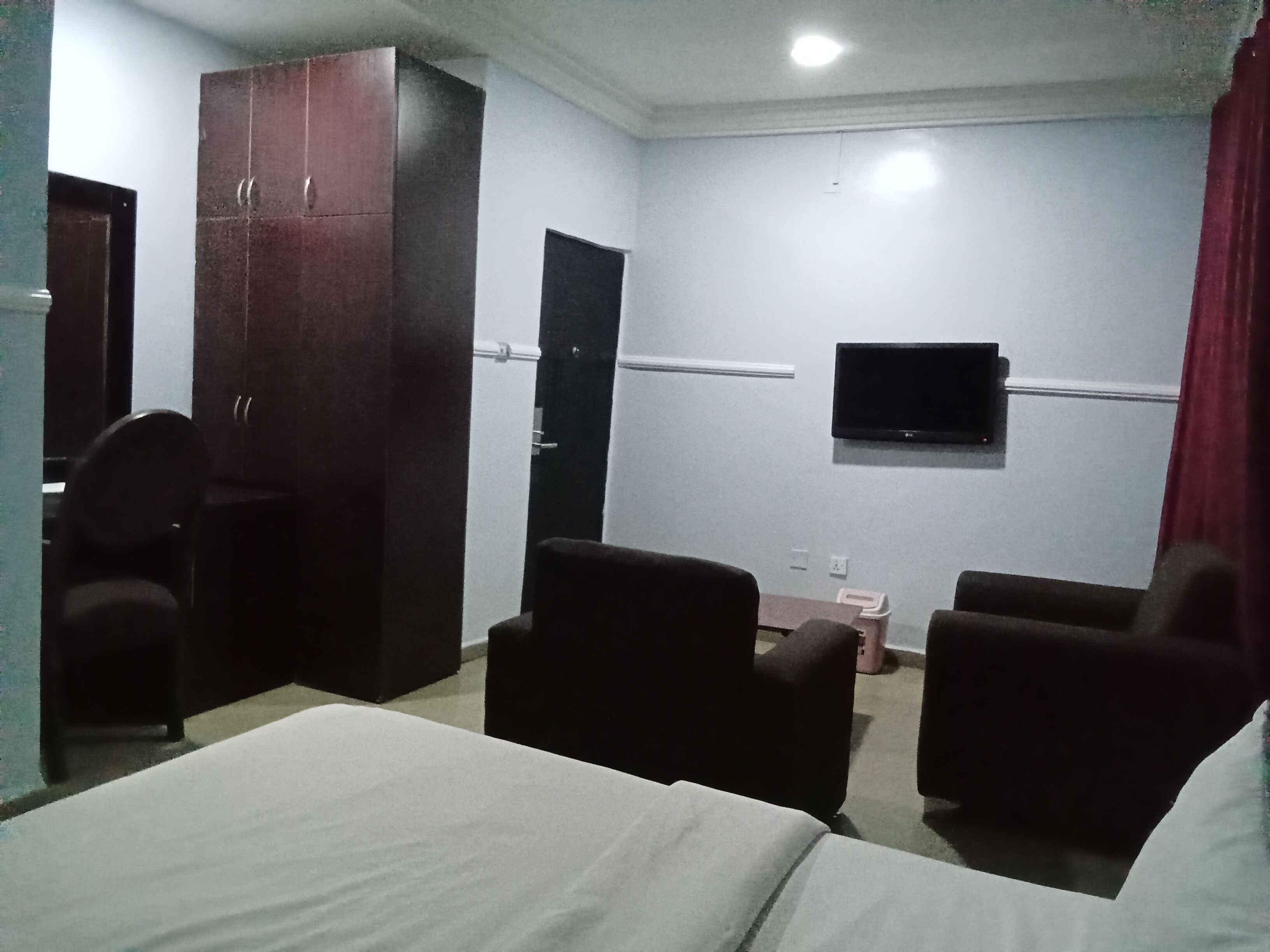 Ein Hotelzimmer mit Bett, zwei Sesseln, einem Kleiderschrank, einem an der Wand befestigten Fernseher und einer Tür.