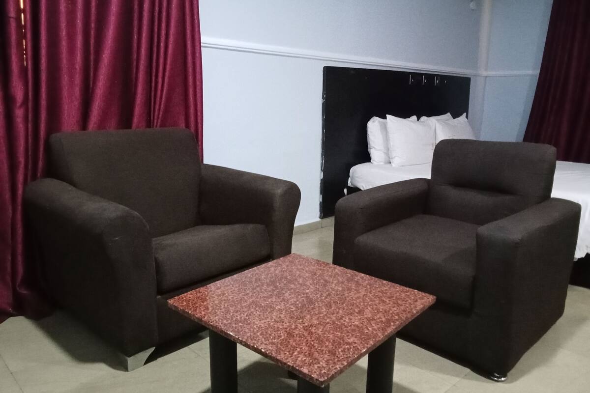 A hotel room with two dark armchairs, a small table, a bed with white bedding, and red curtains.