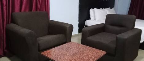 Quarto de hotel com duas poltronas escuras, uma mesa pequena, cama com roupa de cama branca e cortinas vermelhas.