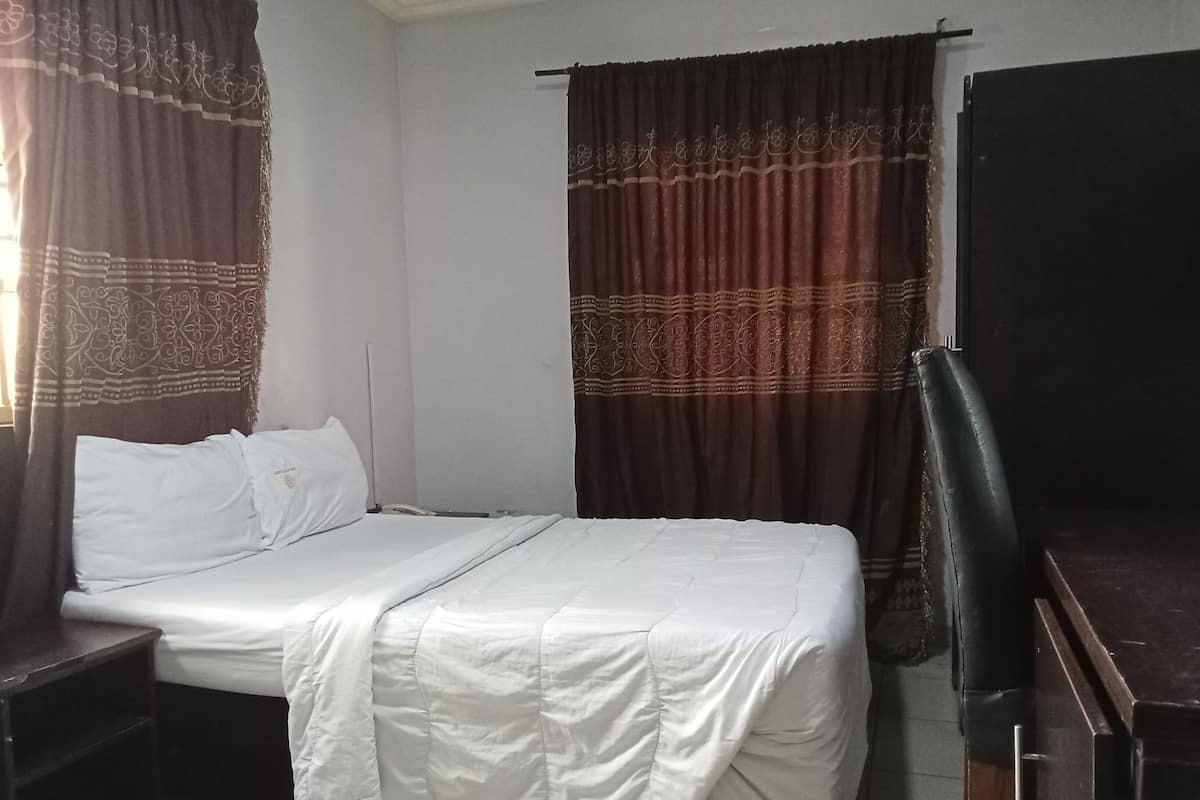 A single bed with a white bedspread, a bedside table, a chair, and a wardrobe.