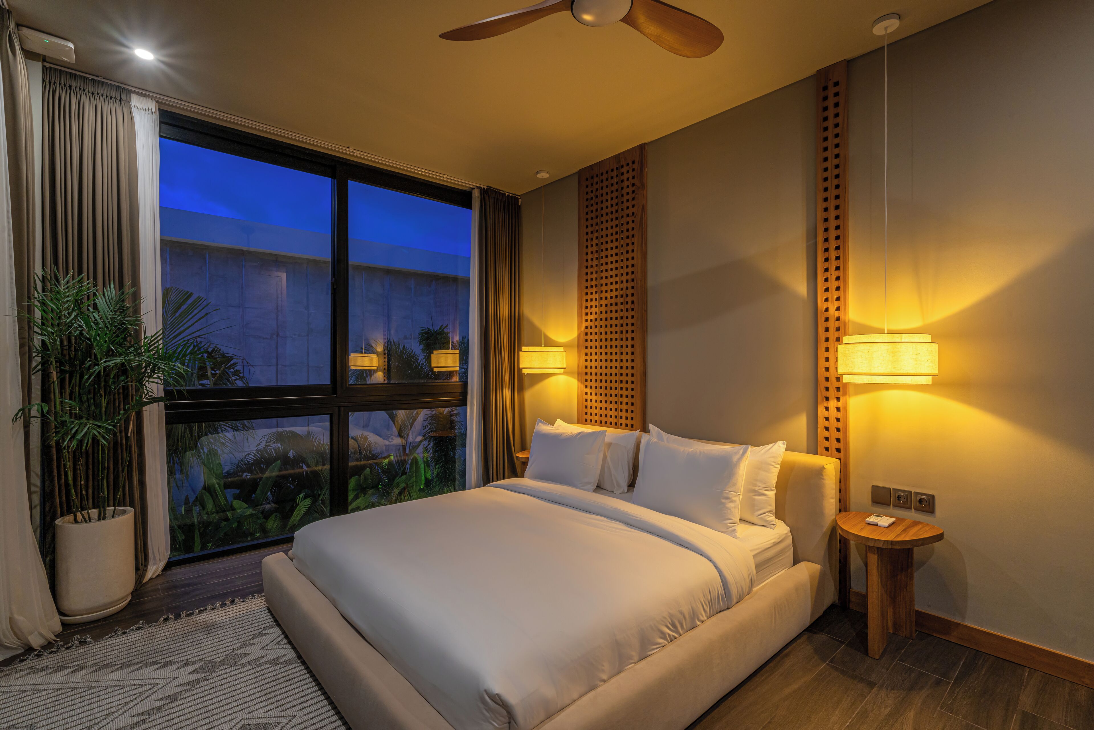 A modern hotel room with a large bed, a ceiling fan, a nightstand, a potted plant, and a view of the outdoors.