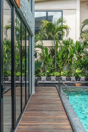 A poolside area with a wooden deck, a seating area with cushions, and lush tropical plants. - Villa Amanora (Canggu)