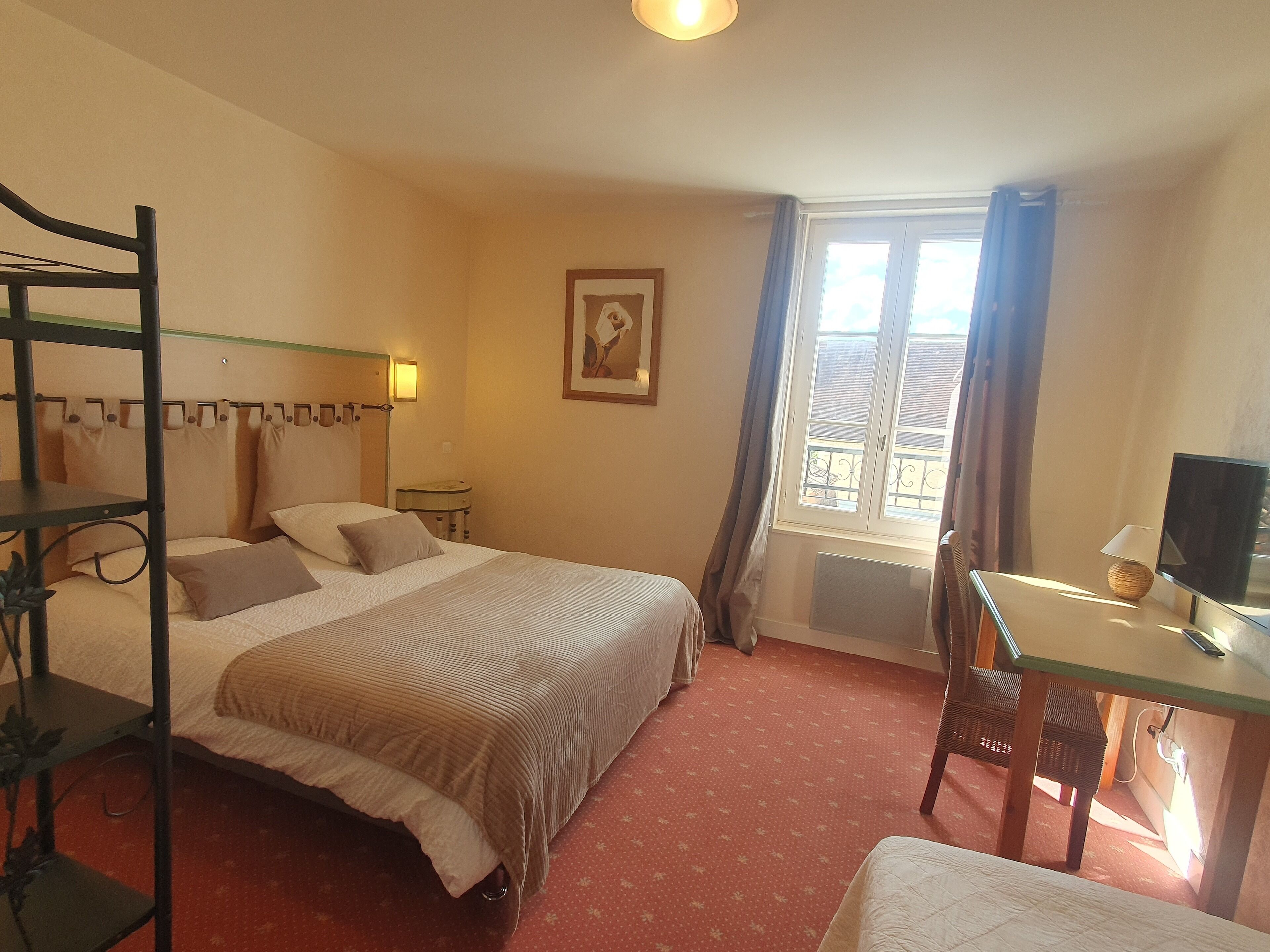 A hotel room with a bed, a desk, a chair, a window with curtains, a wall-mounted lamp, and a framed picture.