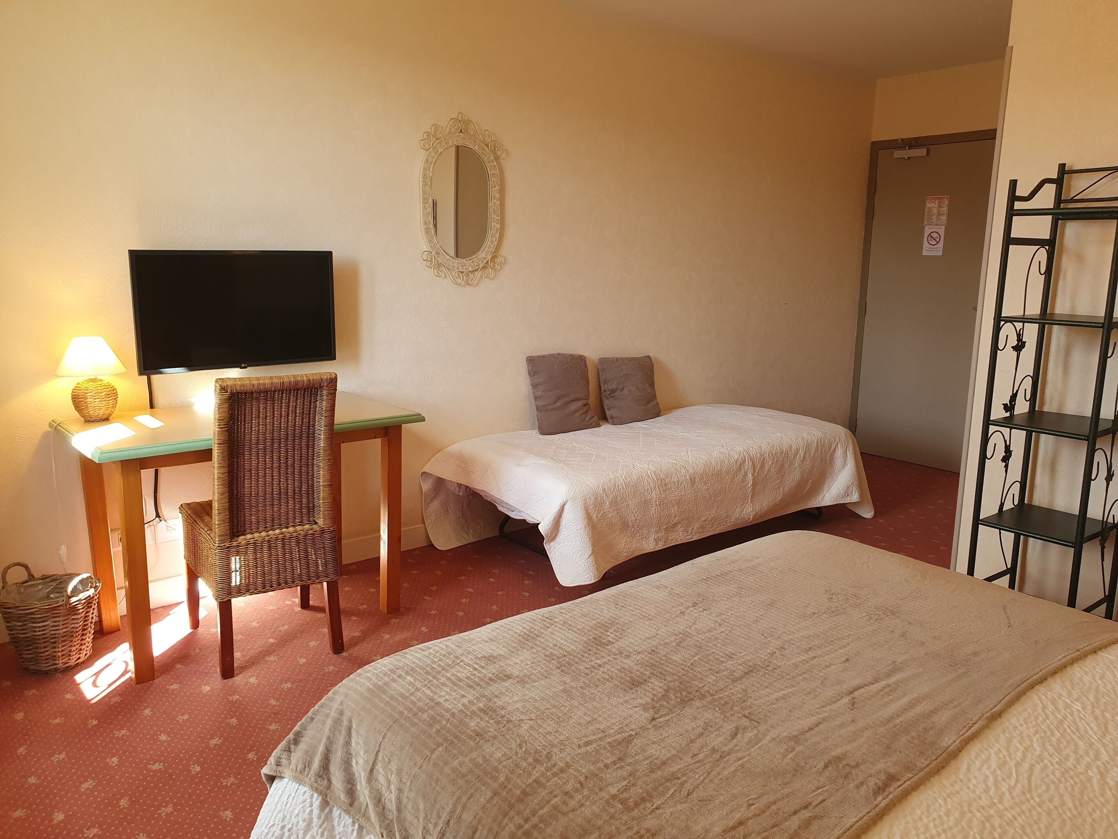 A room with two beds, a desk with a lamp, a television, a mirror, and a metal shelf unit.
