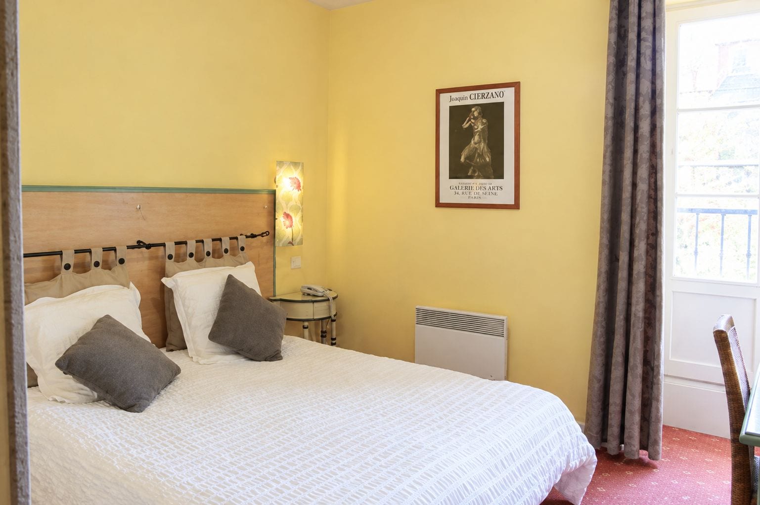 Una habitación de hotel con cama, almohadas, una mesita de noche con lámpara, un radiador y un cartel en la pared.