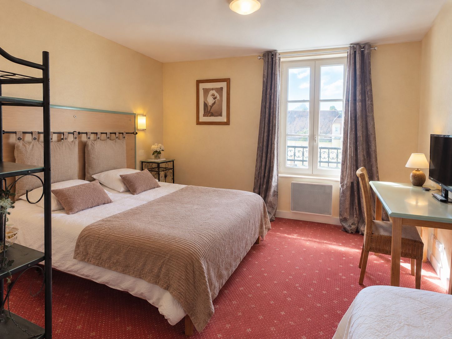 A hotel room with a bed, a desk with a television, a window with curtains, and a wall-mounted picture.