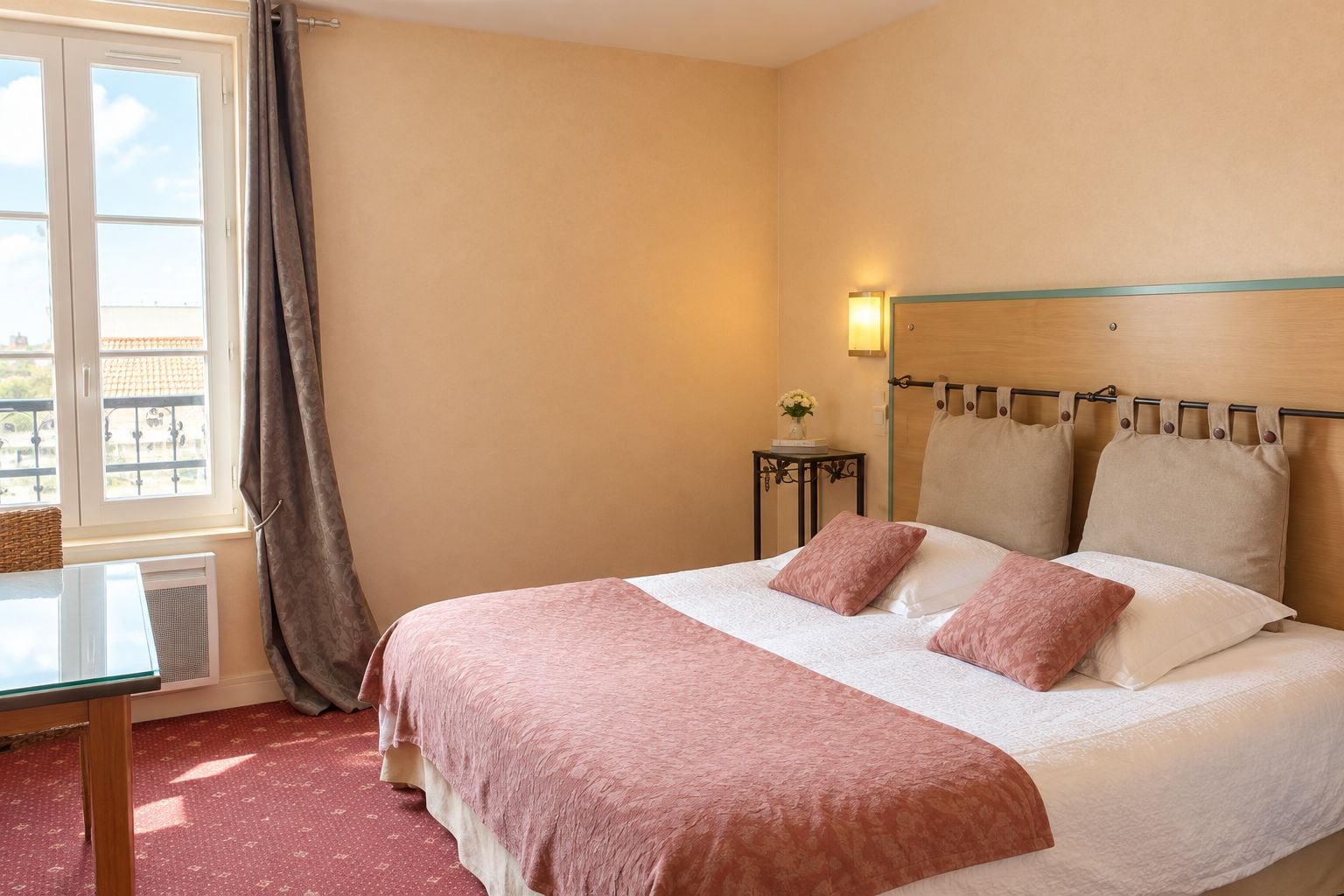 Een hotelkamer met een bed, een tafel, een stoel, een raam met gordijnen en een nachtkastje met een lamp.