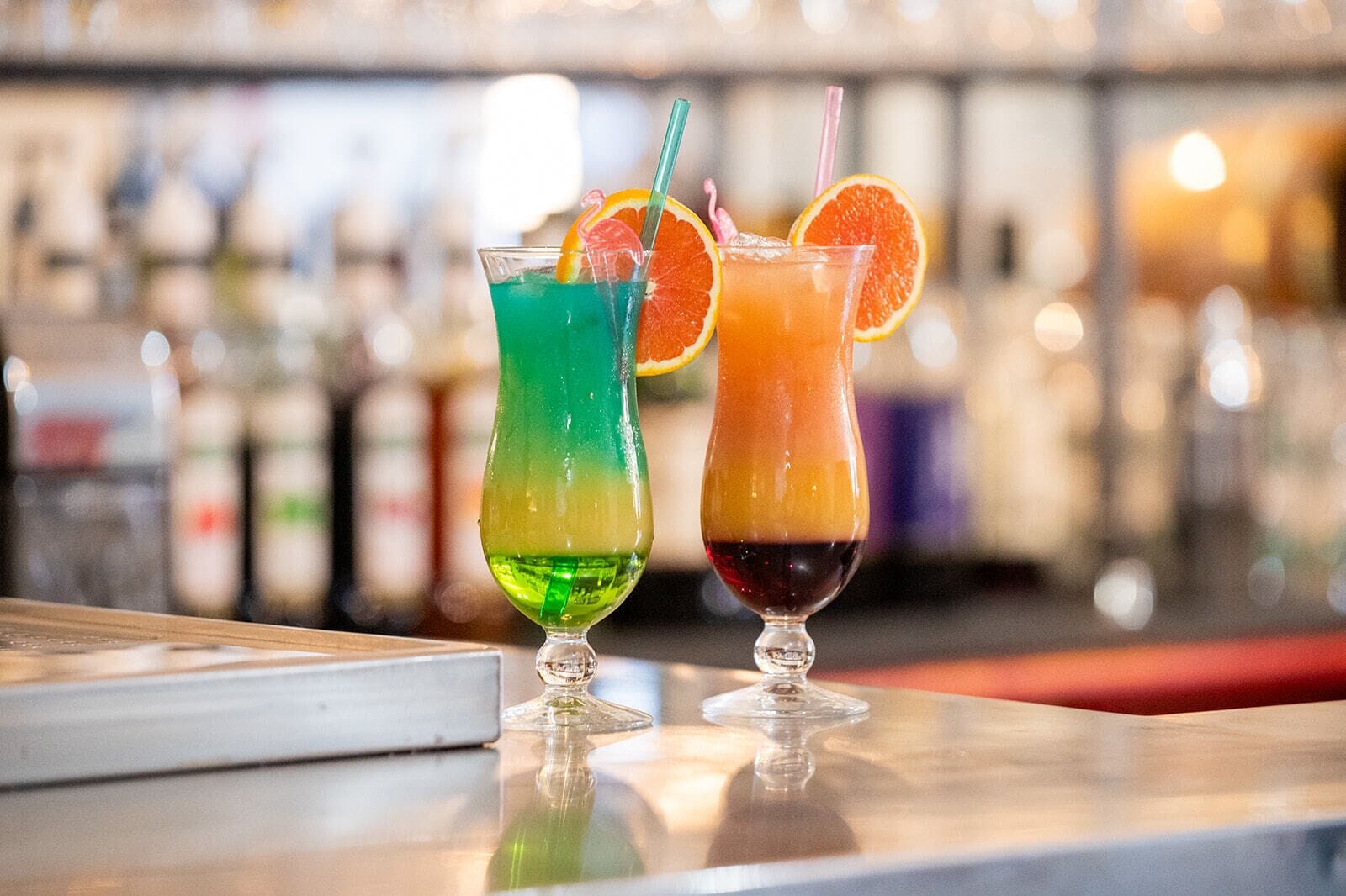 Zwei farbenfrohe Cocktails auf einer Bar, im Hintergrund verschwommene Flaschen.