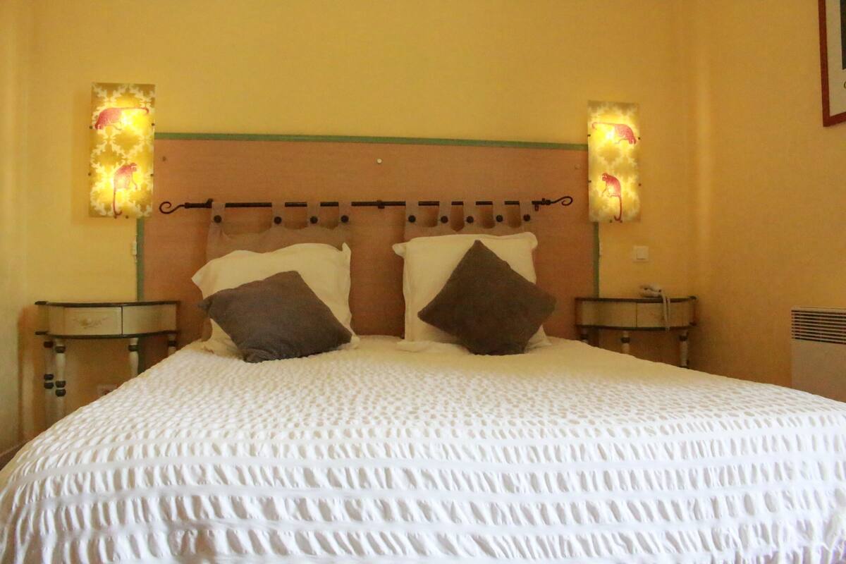 A neatly made bed with a white textured bedspread and two brown pillows. There are two wall-mounted nightstands with lamps on either side of the bed.