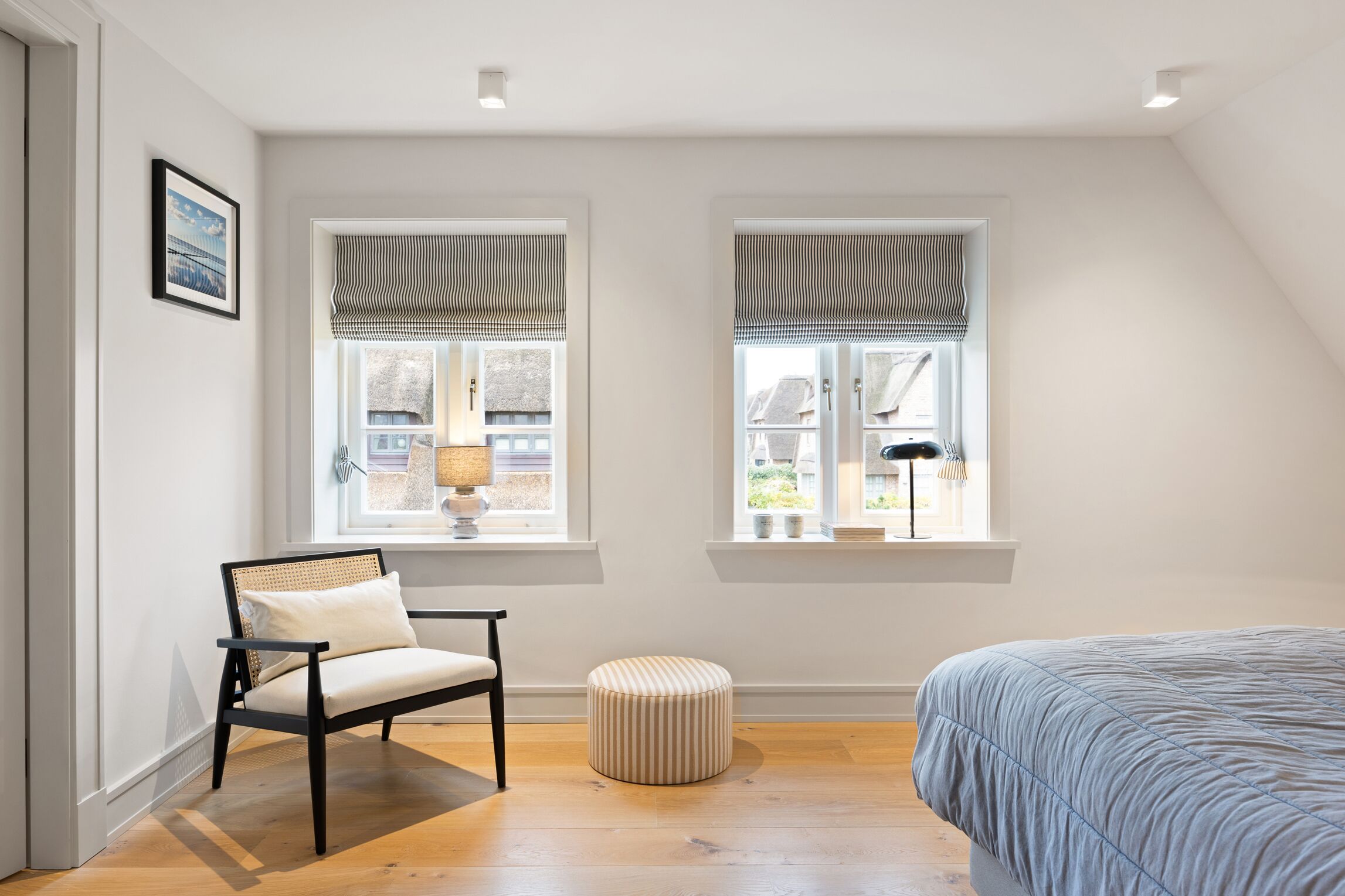 Une chambre à coucher comprenant un lit, une chaise, un petit pouf, deux fenêtres avec des stores et un tableau au mur.