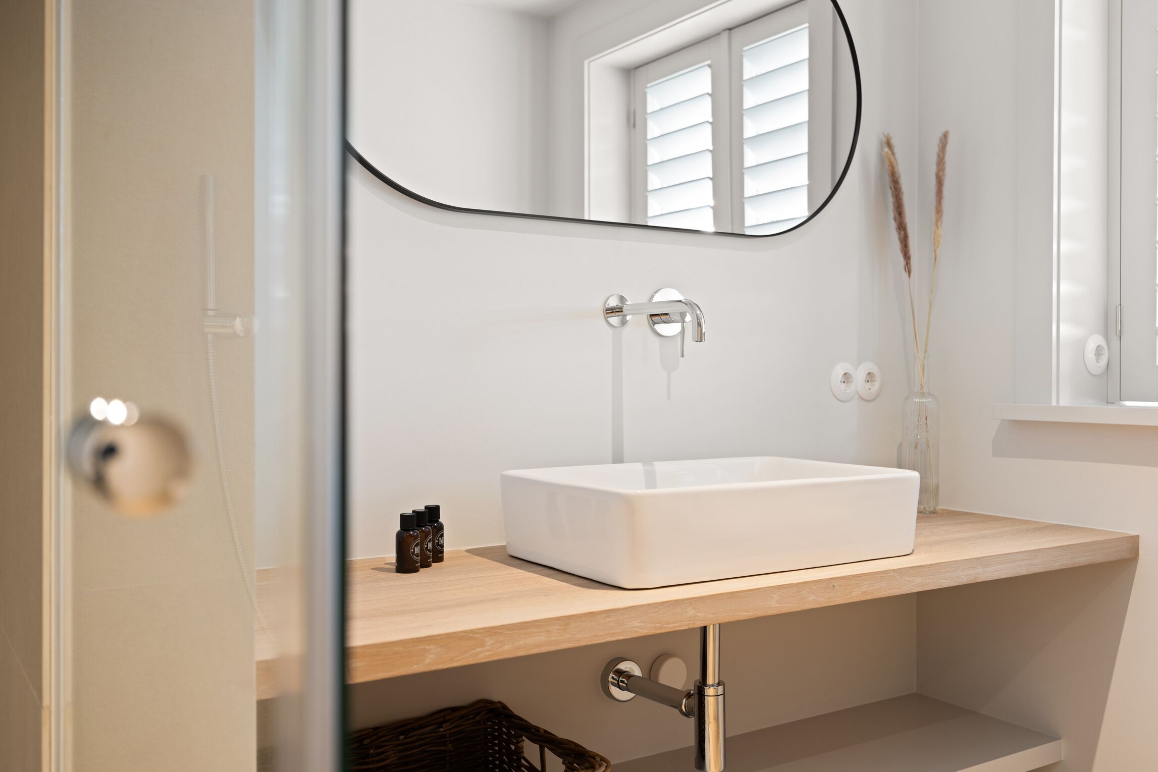A modern bathroom with a white sink, a mirror, and a window with blinds.
