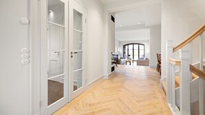 A spacious hallway with a herringbone wooden floor, white walls, and a glass door leading to a room. A staircase with a wooden railing is visible on the right. - Heide.Way - List on Sylt (List auf Sylt)