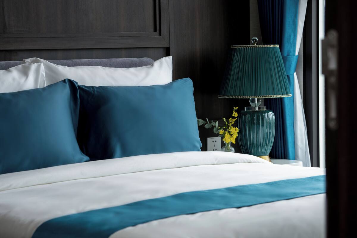 Un lit bien fait, avec une literie bleue et blanche, une lampe turquoise et un vase rempli de fleurs jaunes sur une table de chevet.