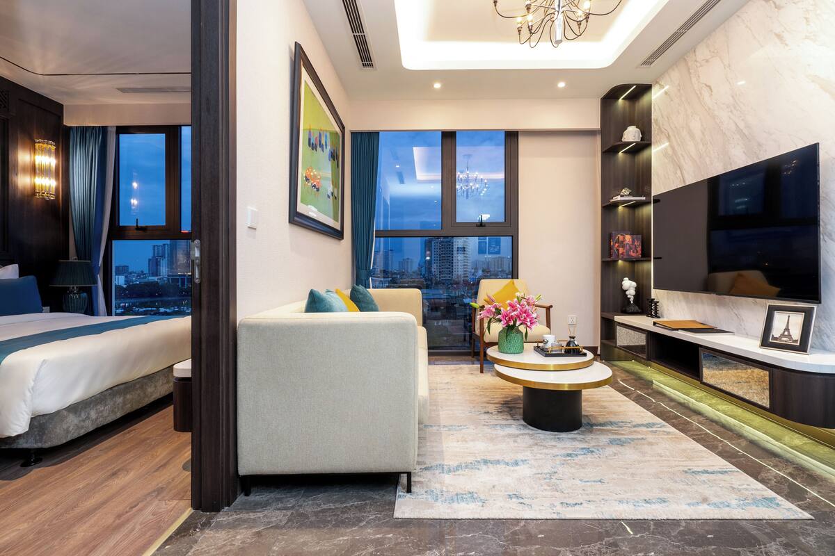 Une chambre d’hôtel moderne équipée d’un lit, d’un canapé, d’une table basse, d’une télévision et de grandes fenêtres donnant sur la ville.