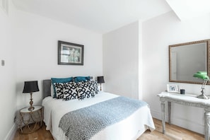 A bedroom with a bed, a bedside table with a lamp, a dresser with a mirror, and a framed picture on the wall.