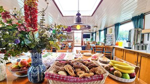 A dining area with a wooden table set with various breads, pastries, and fruits. There is a vase with flowers on the left side of the table.
